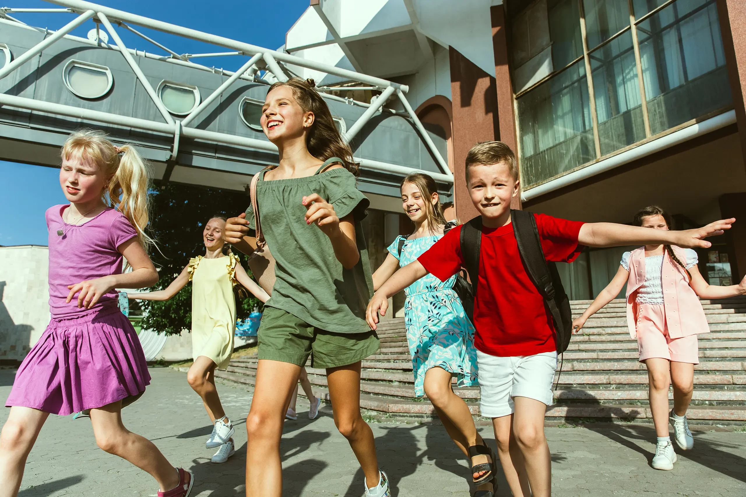 Koffer für Klassenfahrten: Tipps zum Packen und zur Auswahl des Koffers Mehrere fröhliche Kinder laufen lachend in sommerlicher Kleidung und mit Rucksäcken vor einem modernen Schulgebäude
