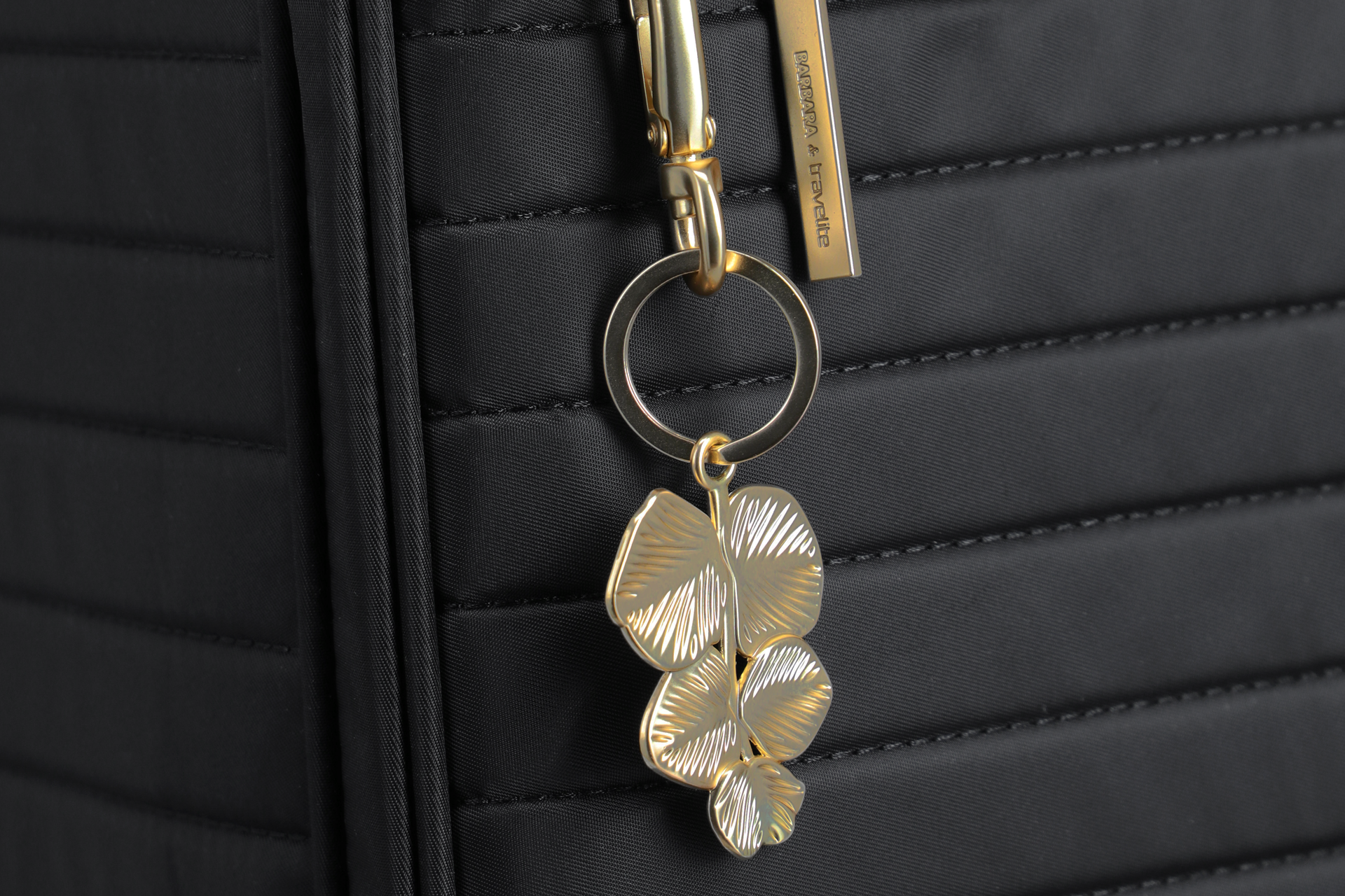 Schwarzer Koffer mit goldfarbenem Schlüsselanhänger und Barbara & Travelite Metallanhänger Detailaufnahme eines schwarzen Koffers mit goldfarbenem Schlüsselanhänger in Form von Blättern und Metallanhänger mit der Gravur Barbara & Travelite
