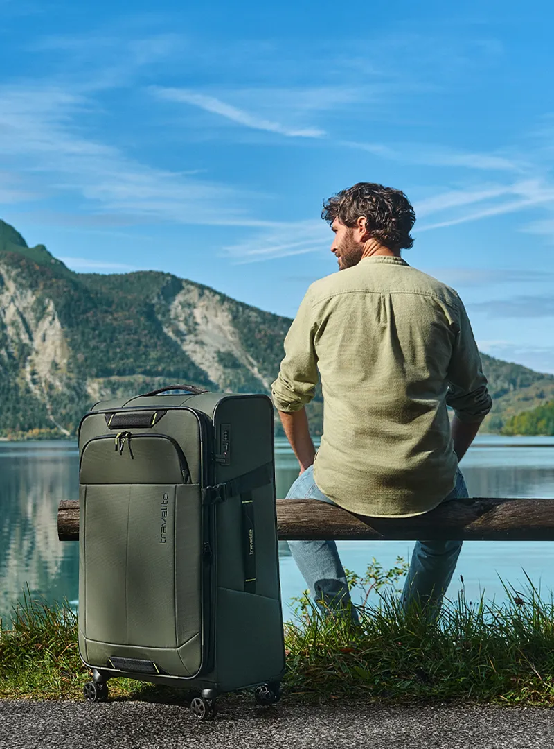 Reisekoffer von Travelite in Naturkulisse – ideal für Ihre nächste Reise Mann sitzt am See mit grünem Travelite Koffer auf Rollen vor Berglandschaft