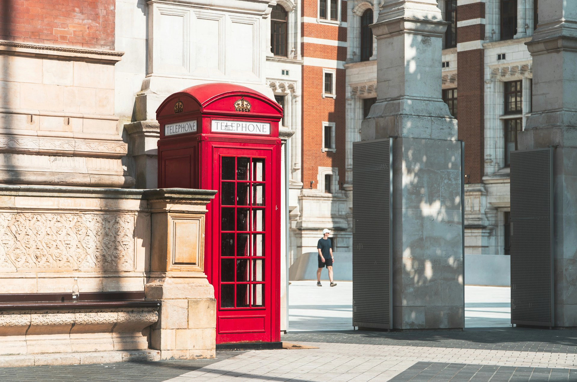A red British telephone box stands on a cobbled square next to an ornate stone building. Other buildings and a man walking between two pillars can be seen in the background. The bright light casts shadows on the square.