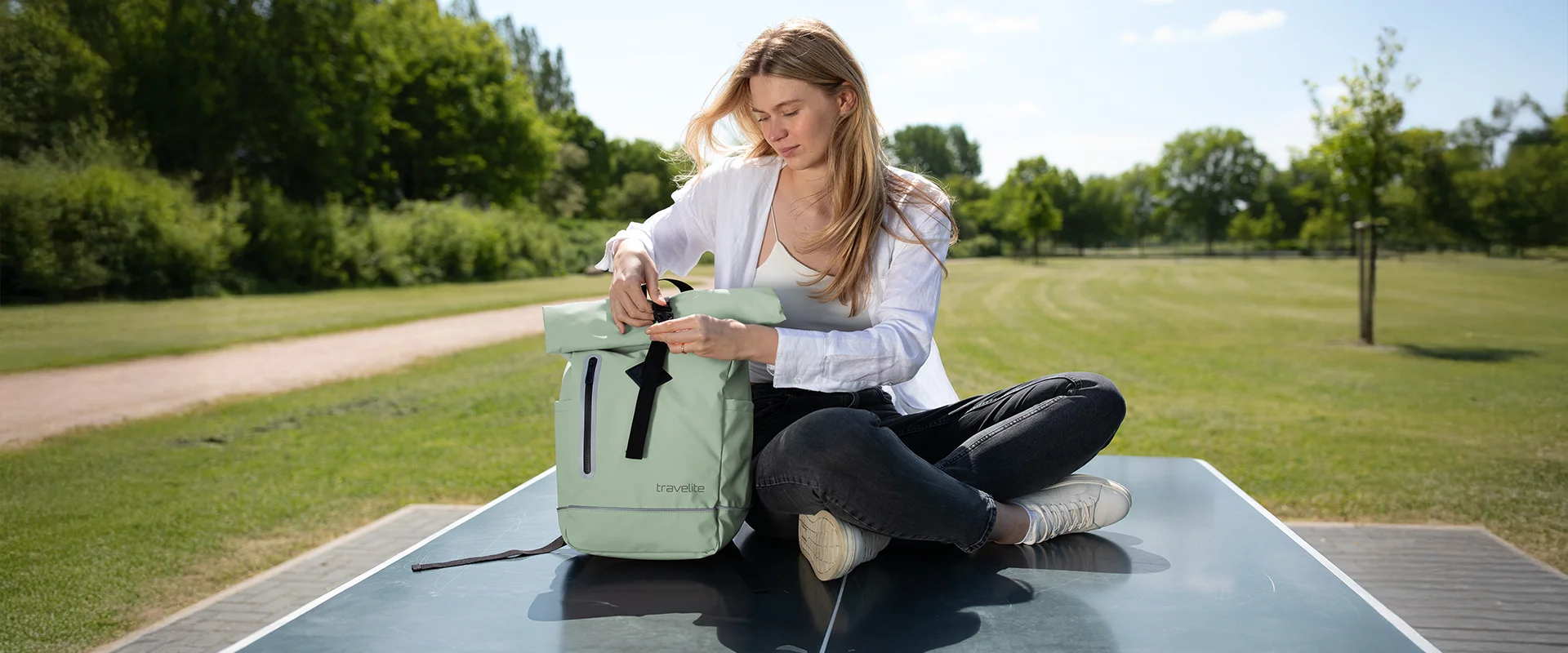 Frau sitzt im Park und öffnet einen mintgrünen travelite Rucksack auf einer Tischtennisplatte Junge Frau sitzt draußen auf einer Tischtennisplatte und öffnet einen mintgrünen travelite Rucksack, sonniges Wetter, grüne Wiese im Hintergrund