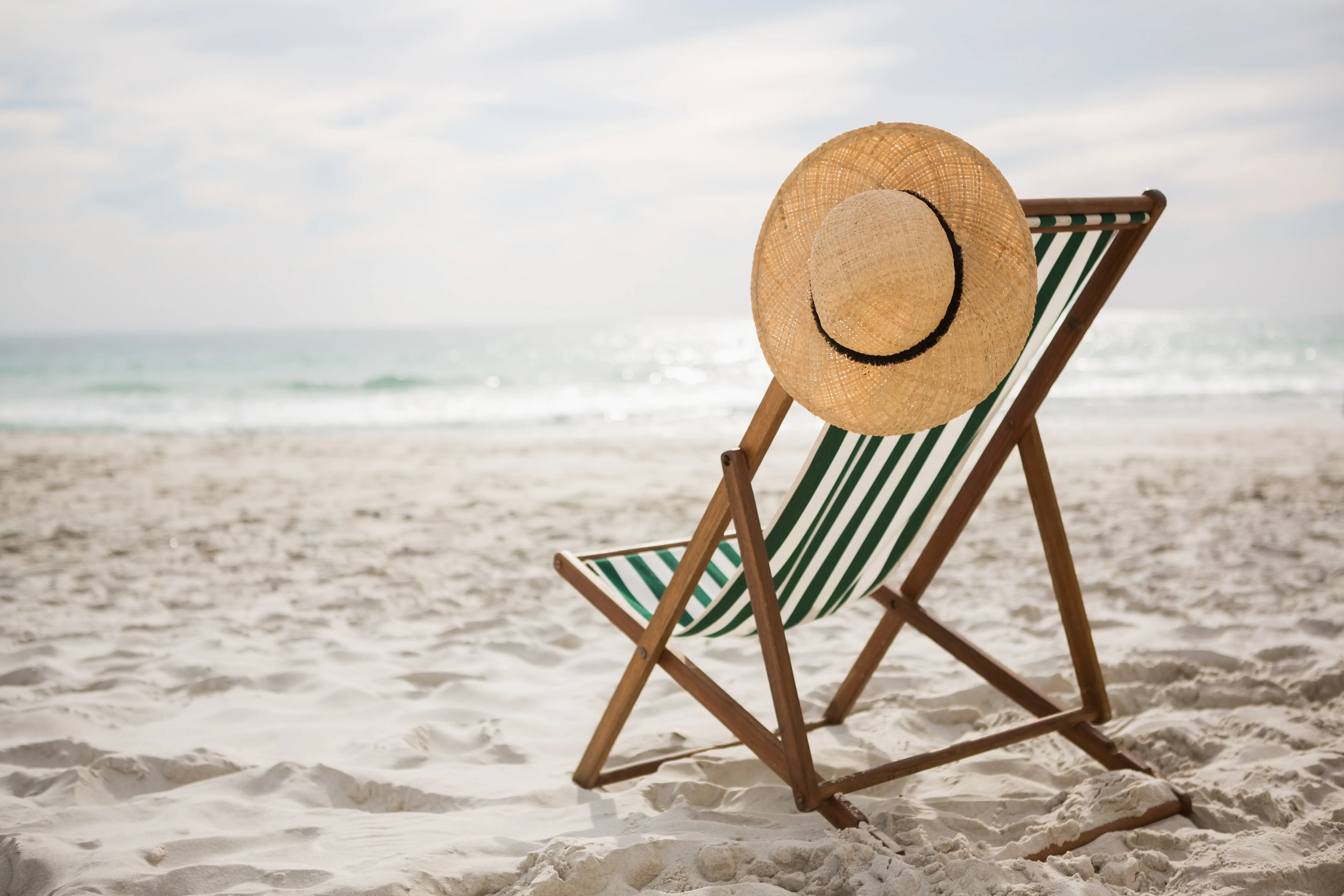 Fernreisen im Sommer – Entdecke 5 Traumziele für unvergessliche Abenteuer Gestreifter Liegestuhl mit Strohhut am sonnigen Sandstrand, im Hintergrund das Meer und blauer Himmel