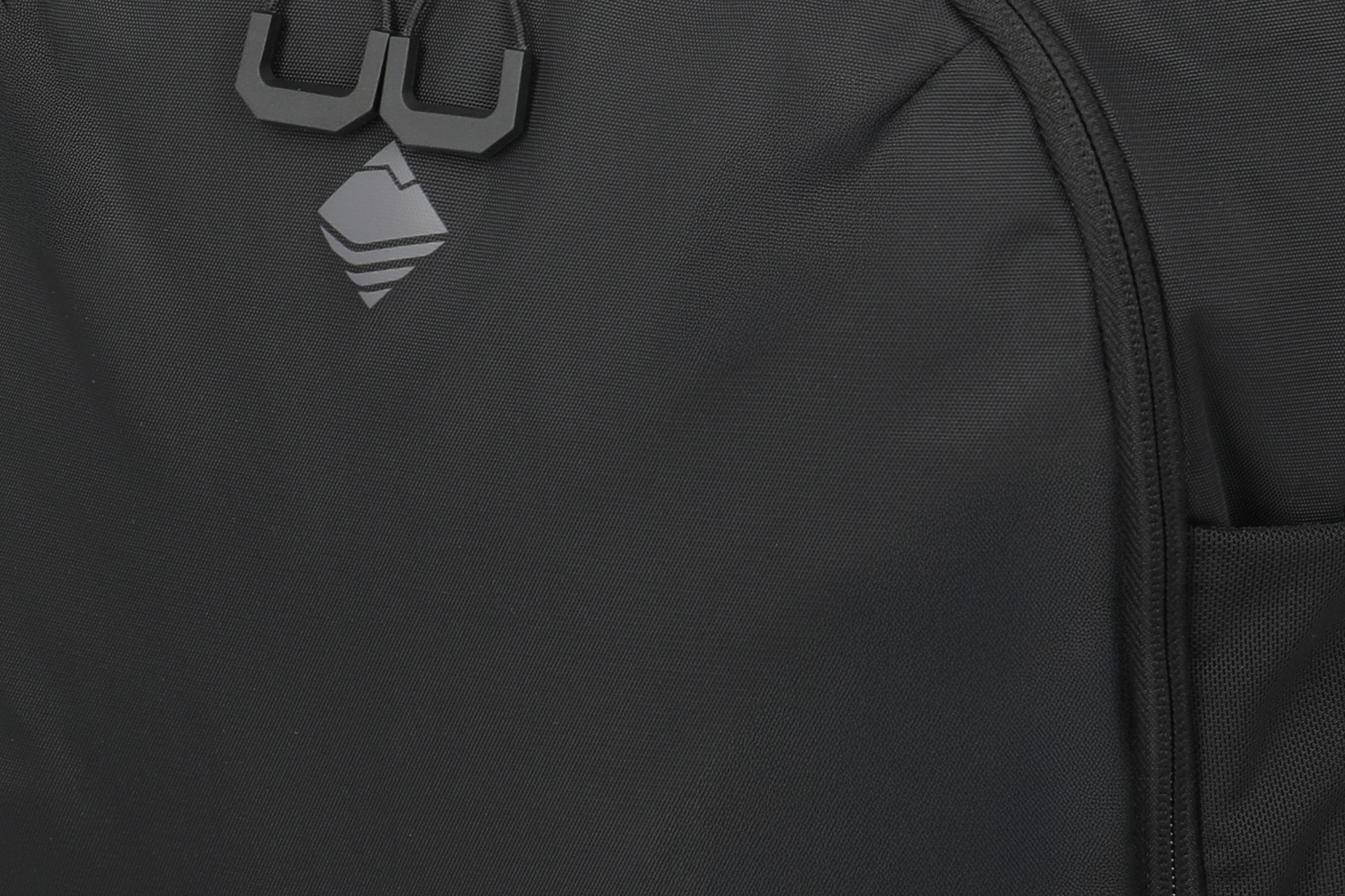 Close-up van een zwarte, licht glanzende rugzakstof. Linksboven zijn twee vierkante ritstrekkers en een grijs, ruitvormig logo met drie horizontale lijnen te zien. Rechts zit een ritssluiting.