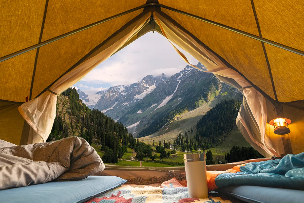Von glamourös bis rustikal: Camping neu entdeckt Aussicht aus einem Zelt auf beeindruckende Berge, grüne Täler, Schlafsäcke und eine Thermoskanne beim Camping