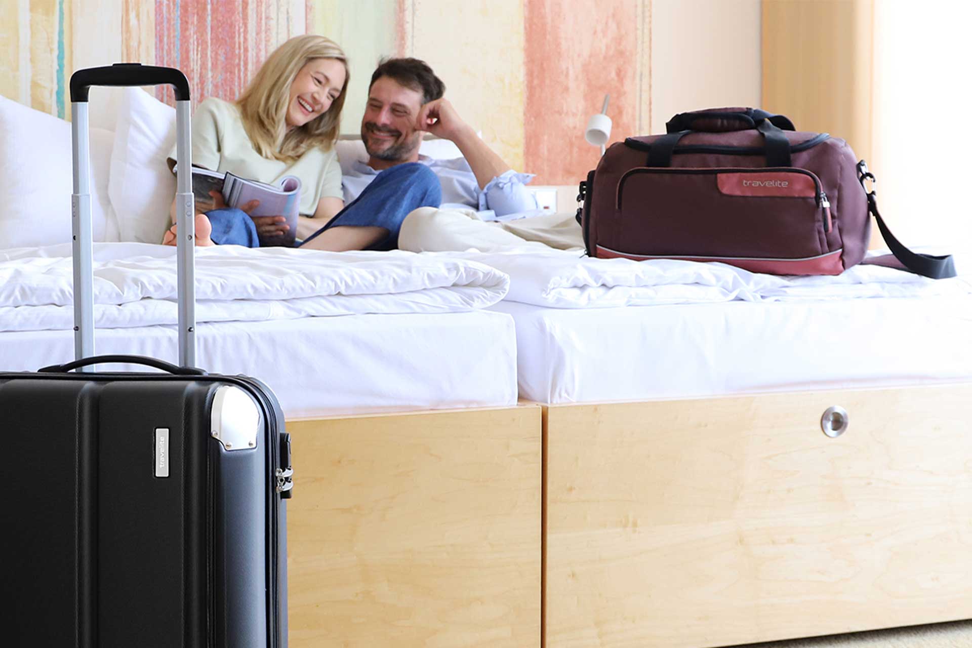 Entspanntes Paar im Hotelzimmer mit Reisekoffern Ein Paar sitzt lächelnd auf einem Bett. Im Vordergrund steht ein schwarzer Hartschalenkoffer, daneben eine braune Reisetasche.