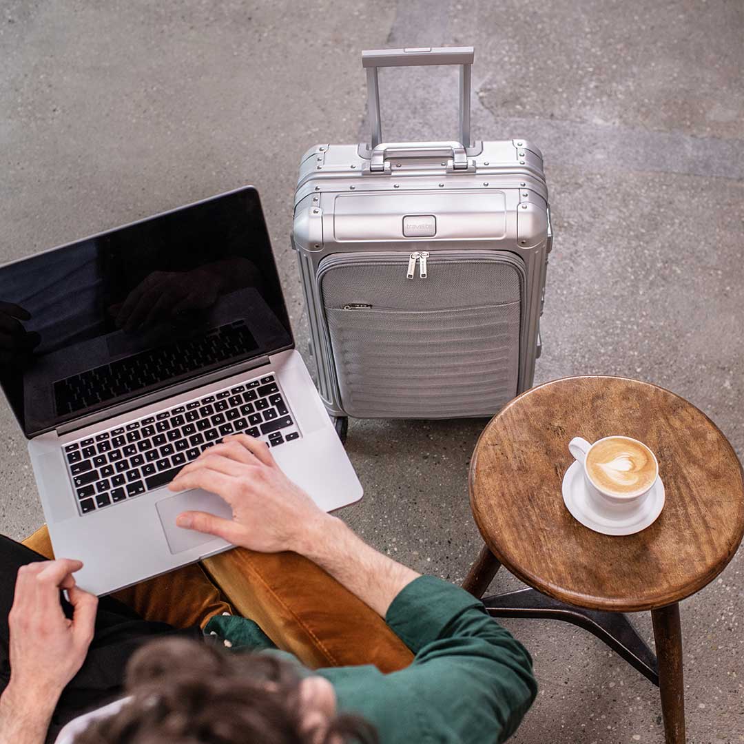 Gepäck Eurowings: das musst du für eine stressfreie Reise wissen Person arbeitet an einem Laptop neben einem silbernen Reisekoffer und einem Cappuccino auf einem Holztisch
