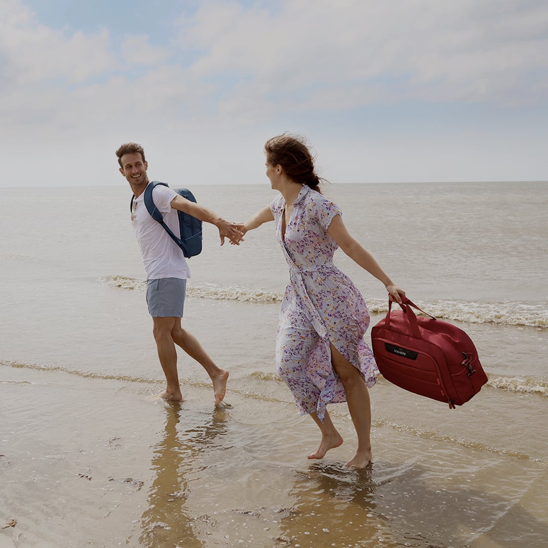 Una coppia si tiene per mano e cammina lungo la spiaggia. L'uomo porta uno zaino, la donna una borsa da viaggio rossa.