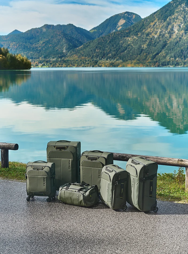 Hochwertige grüne Koffer und Reisetaschen vor malerischer See- und Bergkulisse Mehrere grüne Koffer und Taschen stehen am Ufer eines klaren Sees mit Bergen im Hintergrund bei blauem Himmel