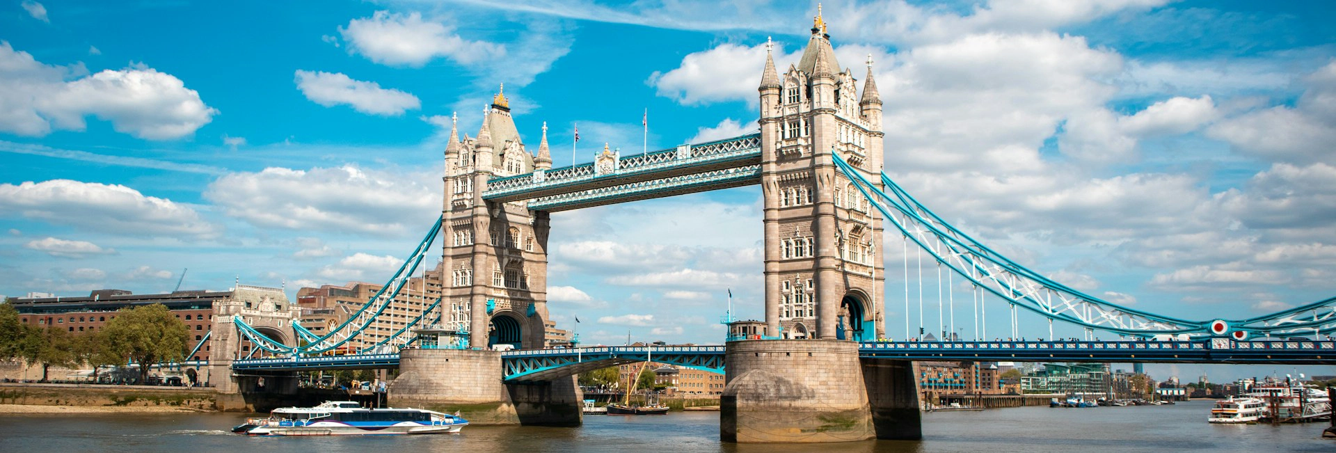 Tower Bridge in London, a stone bridge with two towers, spans a river. Steel cables connect them. Buildings are in the background. The sky is blue with white clouds. A pleasure boat sails on the water.