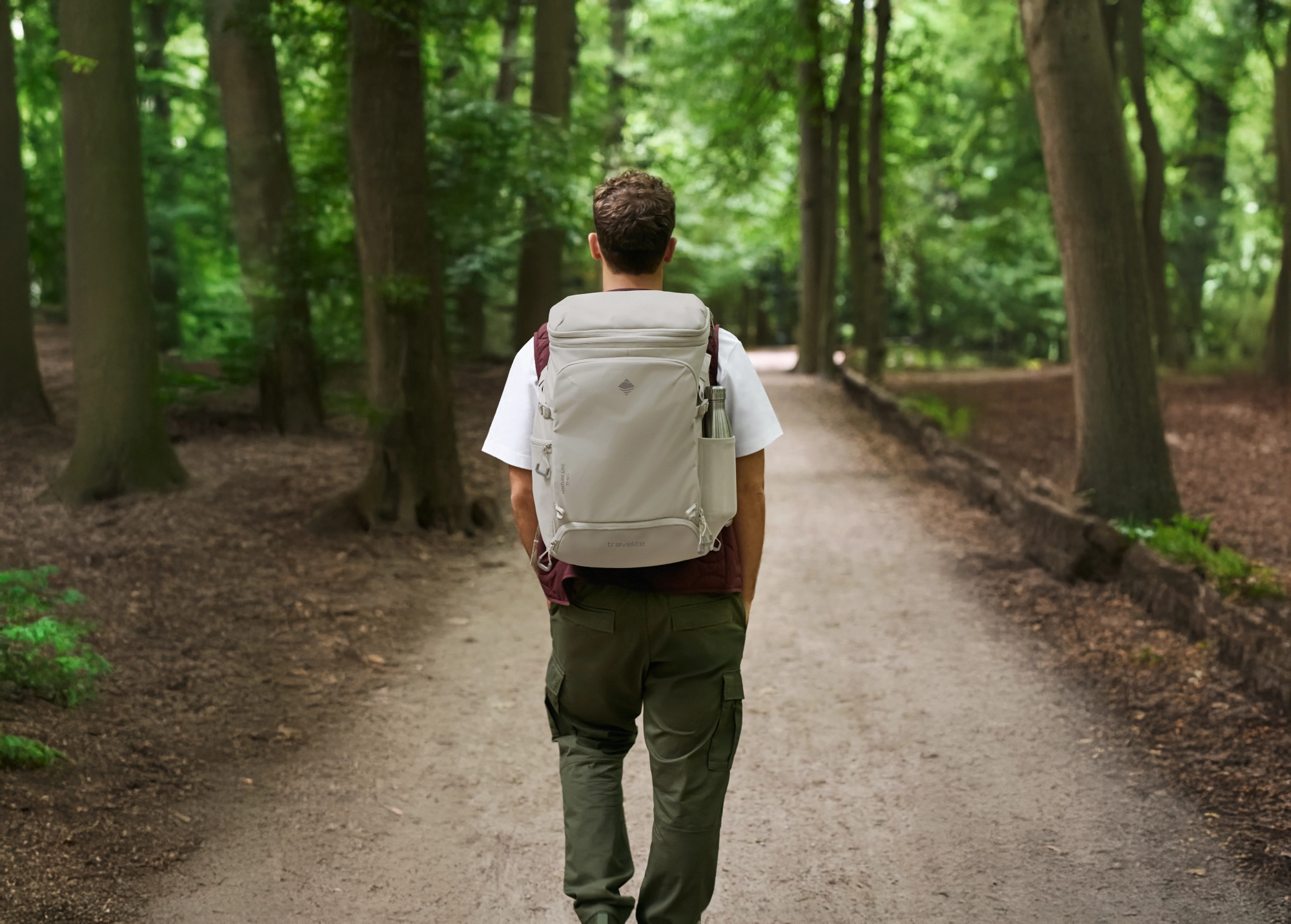 Un hombre de espaldas, con una mochila de color claro en un camino forestal. Lleva una camiseta blanca, un chaleco marrón y pantalones verde oliva. El camino sigue recto, bordeado de árboles. El follaje es denso y verde.