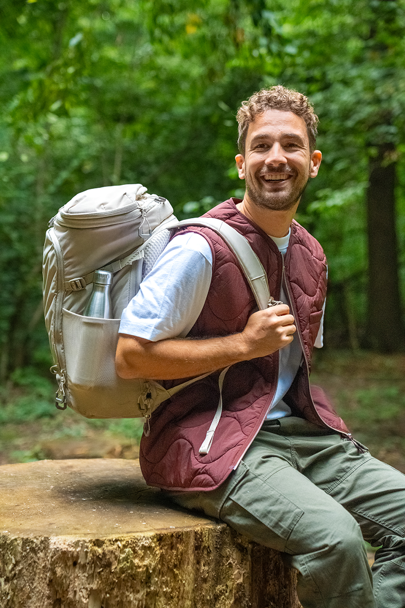 A smiling, light-skinned man is sitting on a tree stump in the forest. He is wearing a burgundy quilted waistcoat over a white T-shirt, green cargo trousers and a light grey rucksack with a water bottle inside.