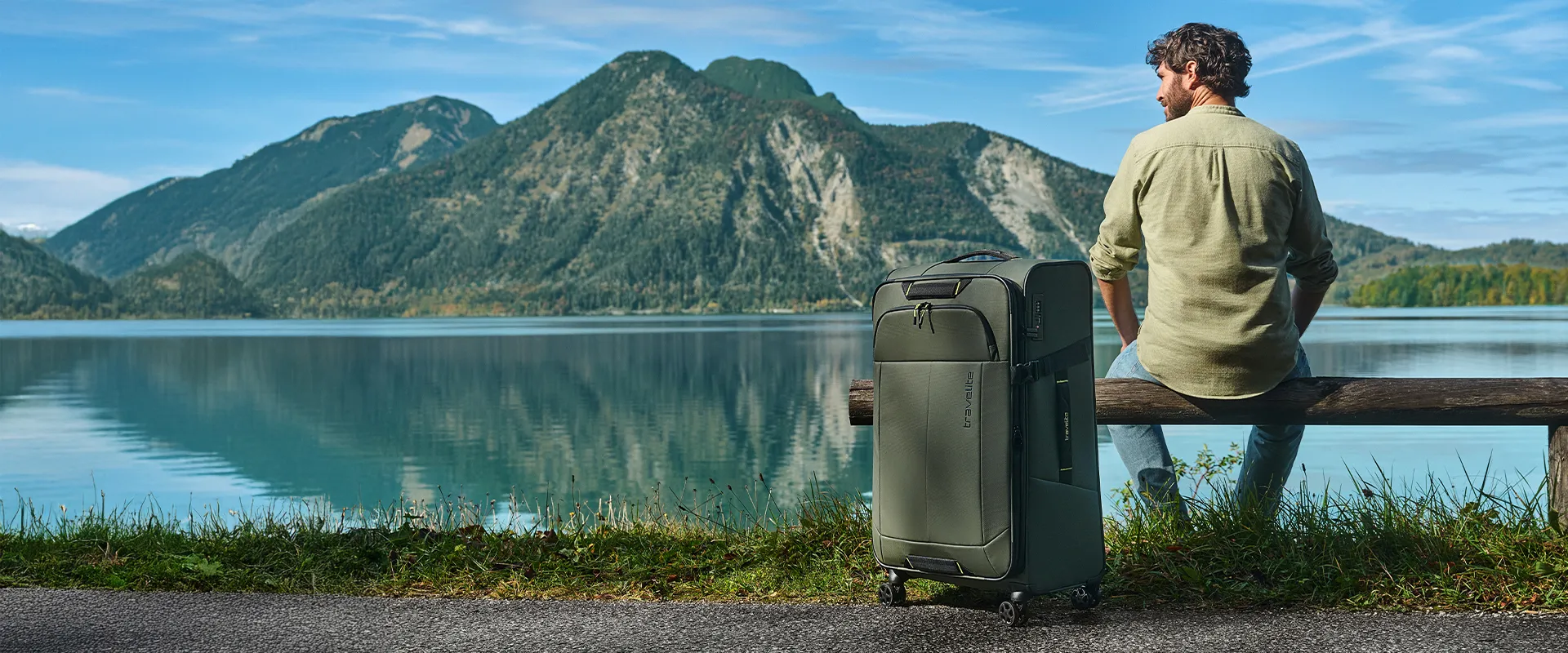 Mann mit grünem Koffer von travelite sitzt an See mit Bergen im Hintergrund Mann sitzt auf Holzbank mit großem, grünen Reisekoffer von travelite am Ufer eines Sees mit Bergen