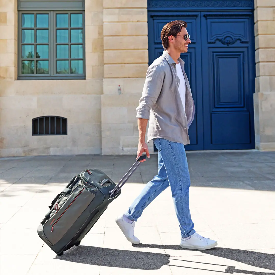 Unterwegs mit der Travelite Rollreisetasche – perfekter Begleiter für den Kurztrip Junger Mann in lässiger Kleidung zieht eine graue Rollreisetasche mit Teleskopgriff vor einem historischen Gebäude entlang
