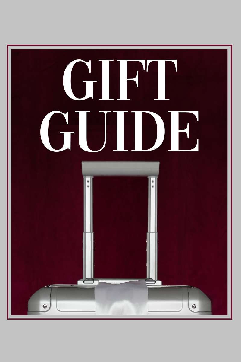 Geschenkideen für Reisen - Stilvolle Kofferwahl Ein eleganter silberner Koffer mit weißer Schleife vor einem tiefroten Hintergrund, begleitet von der Aufschrift 'Gift Guide'.