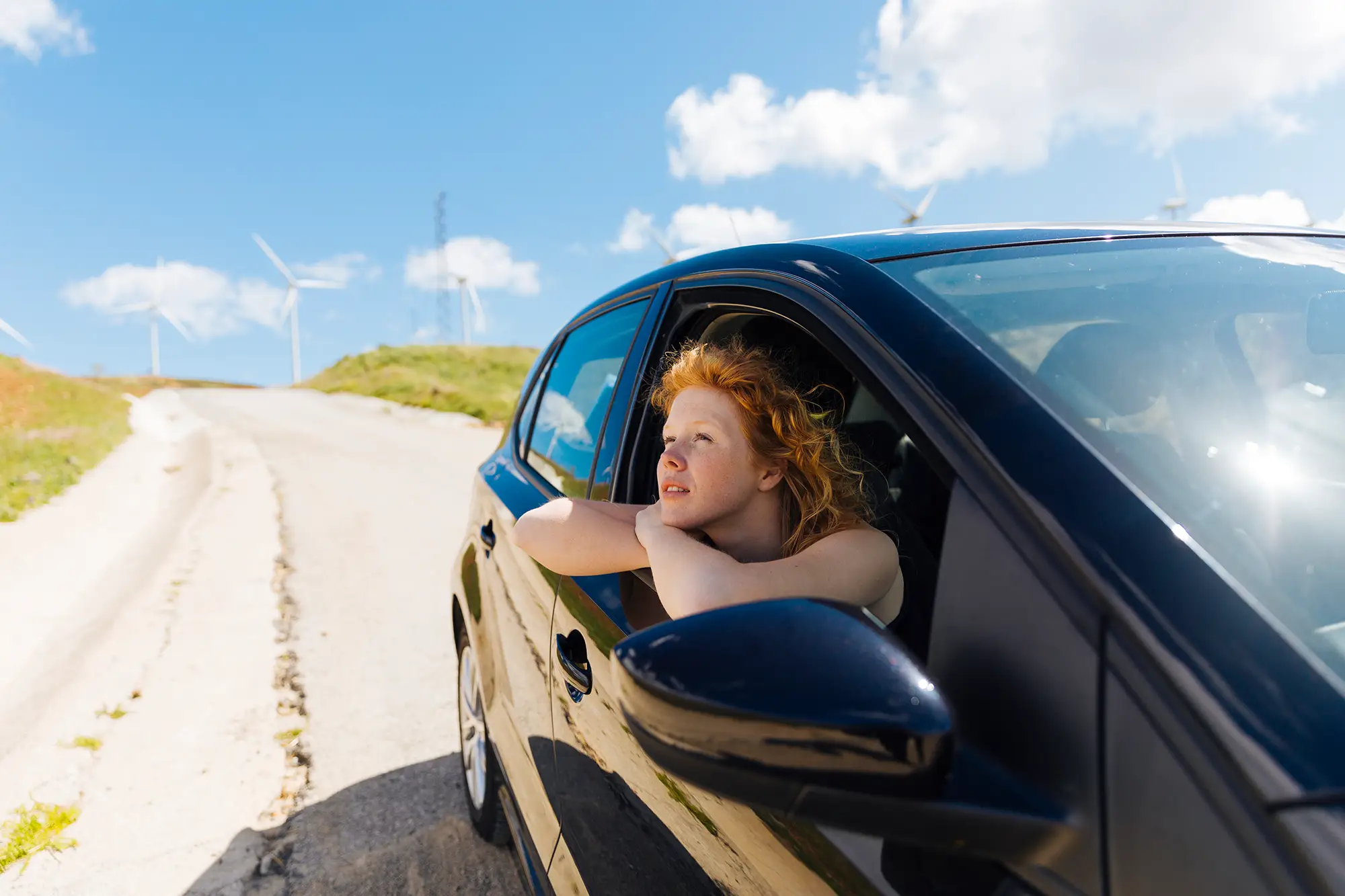 Tipps für lange Autofahrten: So wird deine Fahrt zum Vergnügen Rothaarige Frau lehnt sich aus dem offenen Autofenster und blickt nachdenklich in die Ferne, im Hintergrund Windräder und blauer Himmel