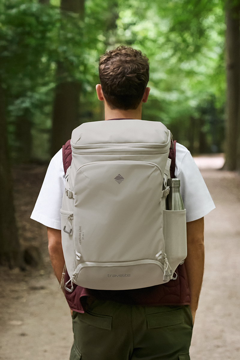 A man with curly brown hair is walking along a forest path. He is wearing a white T-shirt, a wine-red waistcoat and green trousers. On his back is a grey rucksack with "travelite" printed on it. A silver water bottle is attached to his side.
