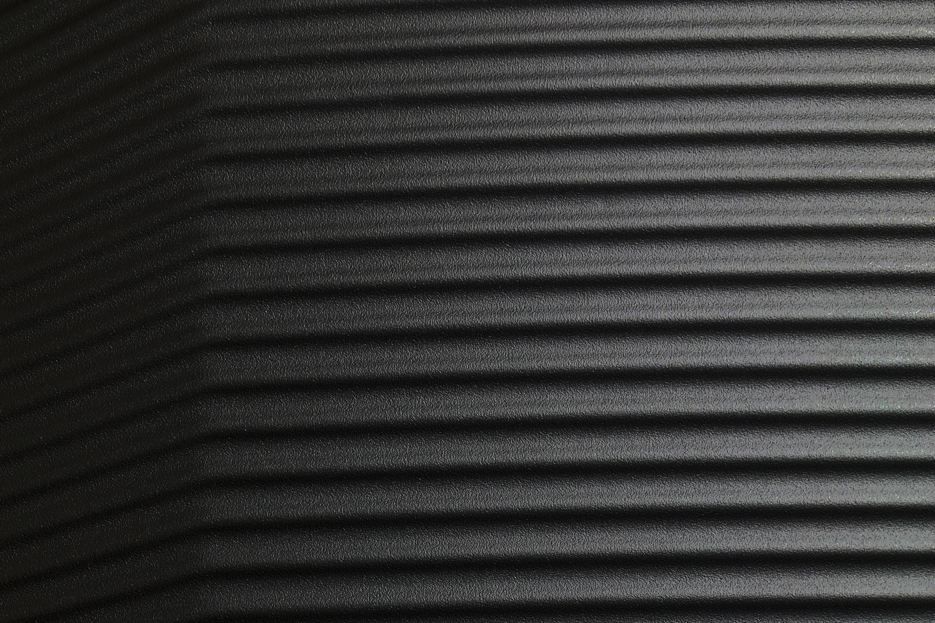 L'immagine mostra un primo piano di una superficie nera scanalata. Le scanalature orizzontali creano un motivo regolare. Un'area a sinistra è più scura. La texture appare ruvida e leggermente lucida.