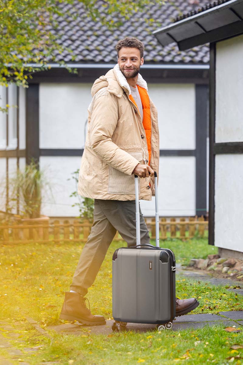 Mann mit beiger Jacke und grauem Rollkoffer im Freien Ein Mann in beiger Winterjacke und orangenem Pullover geht mit einem grauen Rollkoffer durch einen Garten in einem ländlichen Wohngebiet.