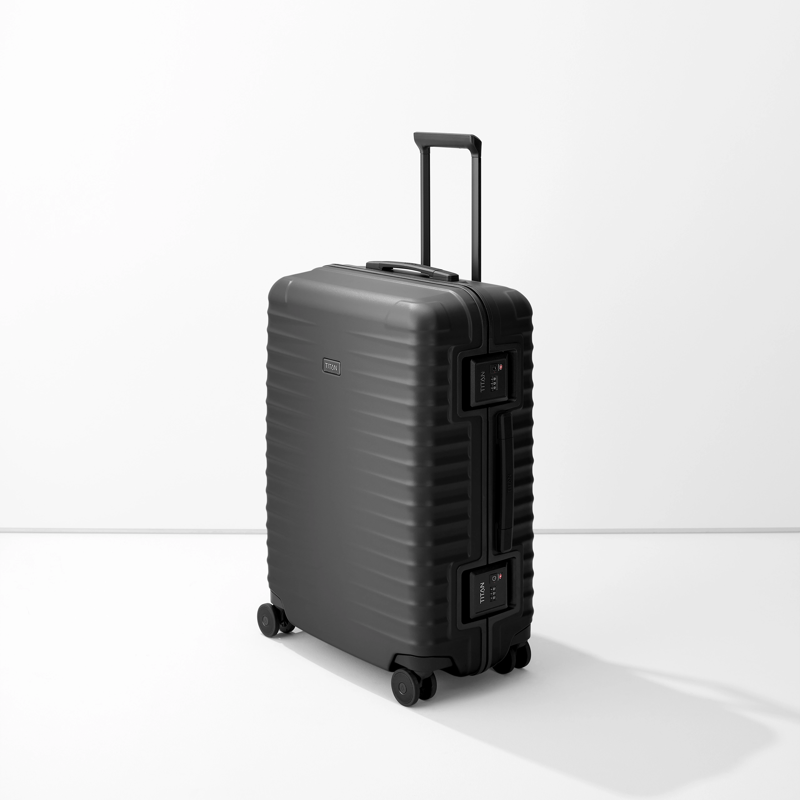 Il trolley nero M+ di TITAN si staglia su uno sfondo bianco. La valigia ha un manico telescopico, una maniglia per il trasporto e due chiusure TSA. La superficie presenta linee orizzontali e il logo "TITAN" è visibile.