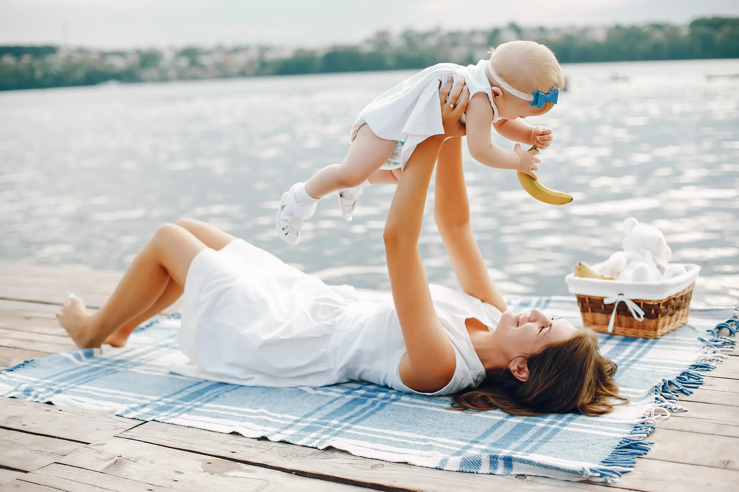 Urlaub mit Baby – Tipps und Tricks für eine stressfreie Reise Frau liegt auf einer Decke am Steg und hebt lachendes Baby hoch, neben ihnen ein Picknickkorb mit Banane und Stofftier vor einem See