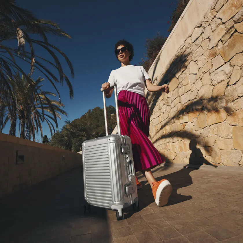Travelite Hartschalenkoffer Silber – eleganter Begleiter für sonnige Reisen Frau in pinkem Rock zieht silbernen Hartschalenkoffer an Steinmauer entlang
