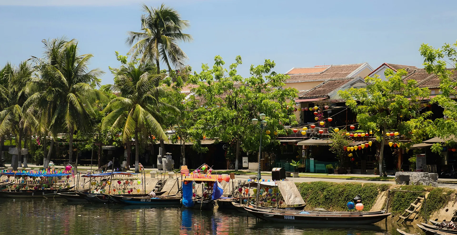 Deine vielfältige Reise durch Vietnam Mehrere Holzboote mit bunten Lampions am Flussufer, im Hintergrund Palmen und traditionelle Häuser in Hoi An, Vietnam