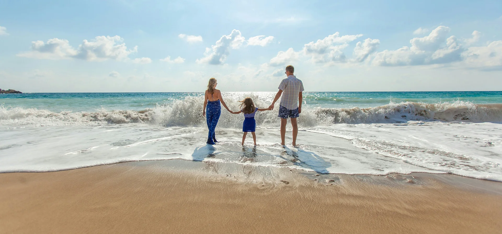 To the sea with the whole family Drei Personen, zwei Erwachsene und ein Kind, stehen Hand in Hand am Strand und blicken auf das Meer mit Wellen und blauem Himmel