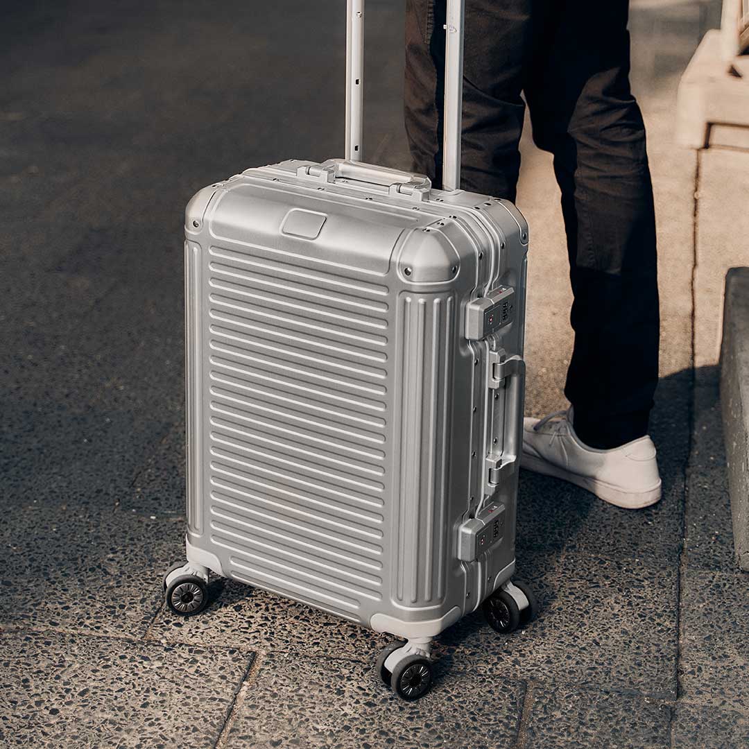 Moderner silberner Koffer mit Rollen auf einem Gehweg Silberner Reisekoffer mit Teleskopgriff und vier Rollen, auf Asphalt stehend neben einer Person in schwarzer Hose und weißen Schuhen