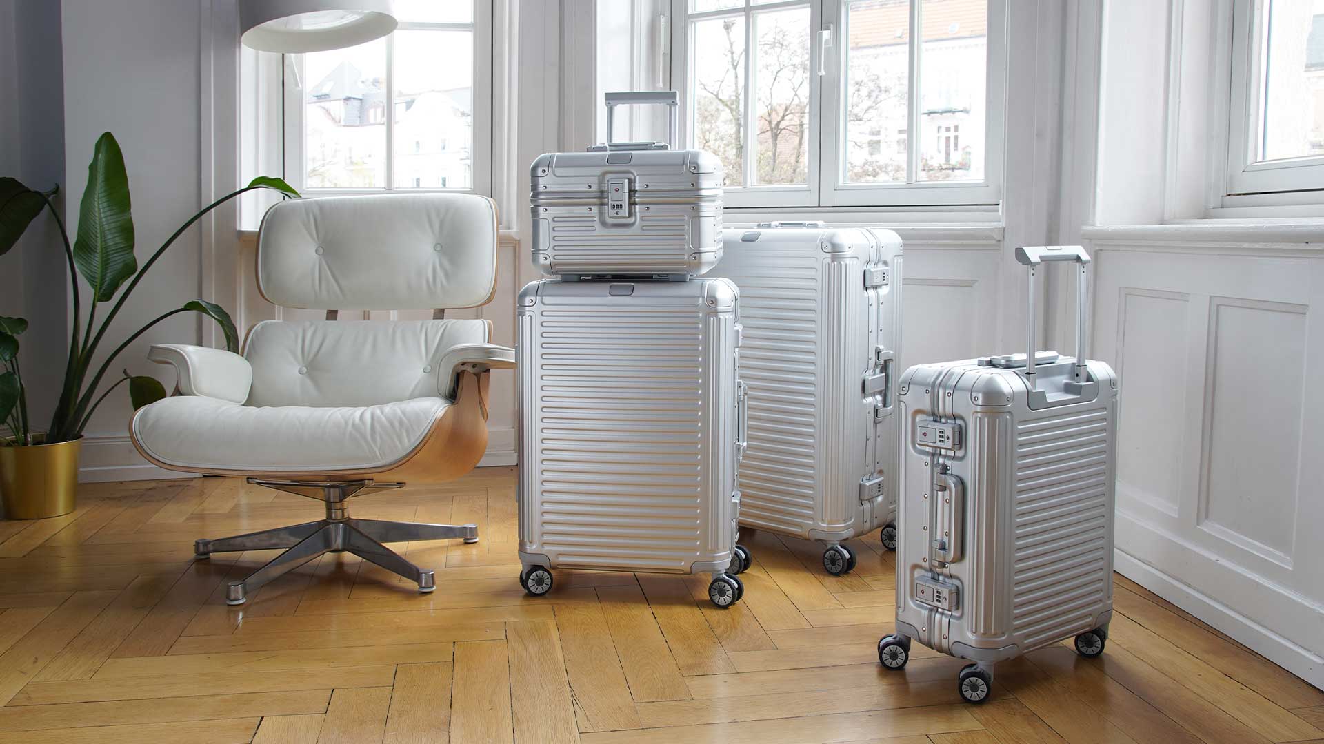 Dimensioni valigie a confronto: trova la misura perfetta per il tuo viaggio con travelite Vier stilvolle Aluminiumkoffer unterschiedlicher Größe neben einem modernen weißen Sessel in einem hellen Raum mit hölzernem Boden und großen Fenstern.