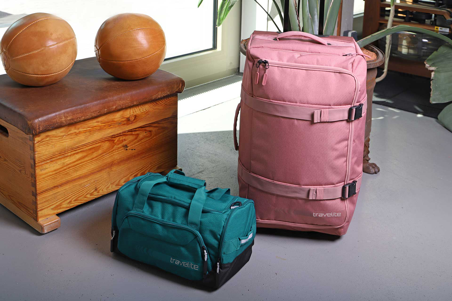 Travelite Reisetaschen-Set in Pink und Türkis neben Turnbank mit Leder-Medizinbällen Zwei Reisetaschen von Travelite in Pink und Türkis stehen auf dem Boden neben einer Holz-Turnbank mit Leder-Medizinbällen