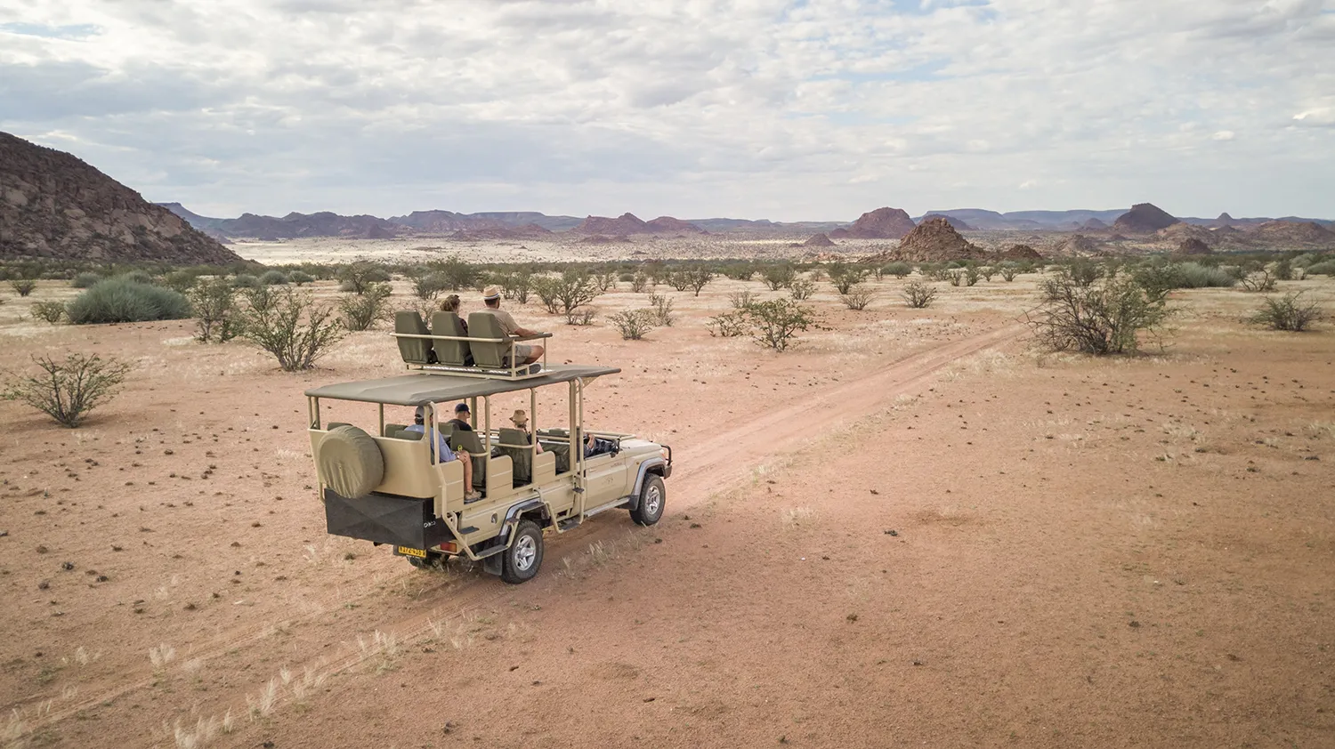 Safari durch Namibia: So gelingt Dein Roadtrip Gruppe von Menschen auf Safari-Fahrt im offenen Geländewagen durch eine trockene, weite Wüstenlandschaft mit vereinzelten Büschen und Bergen im Hintergrund