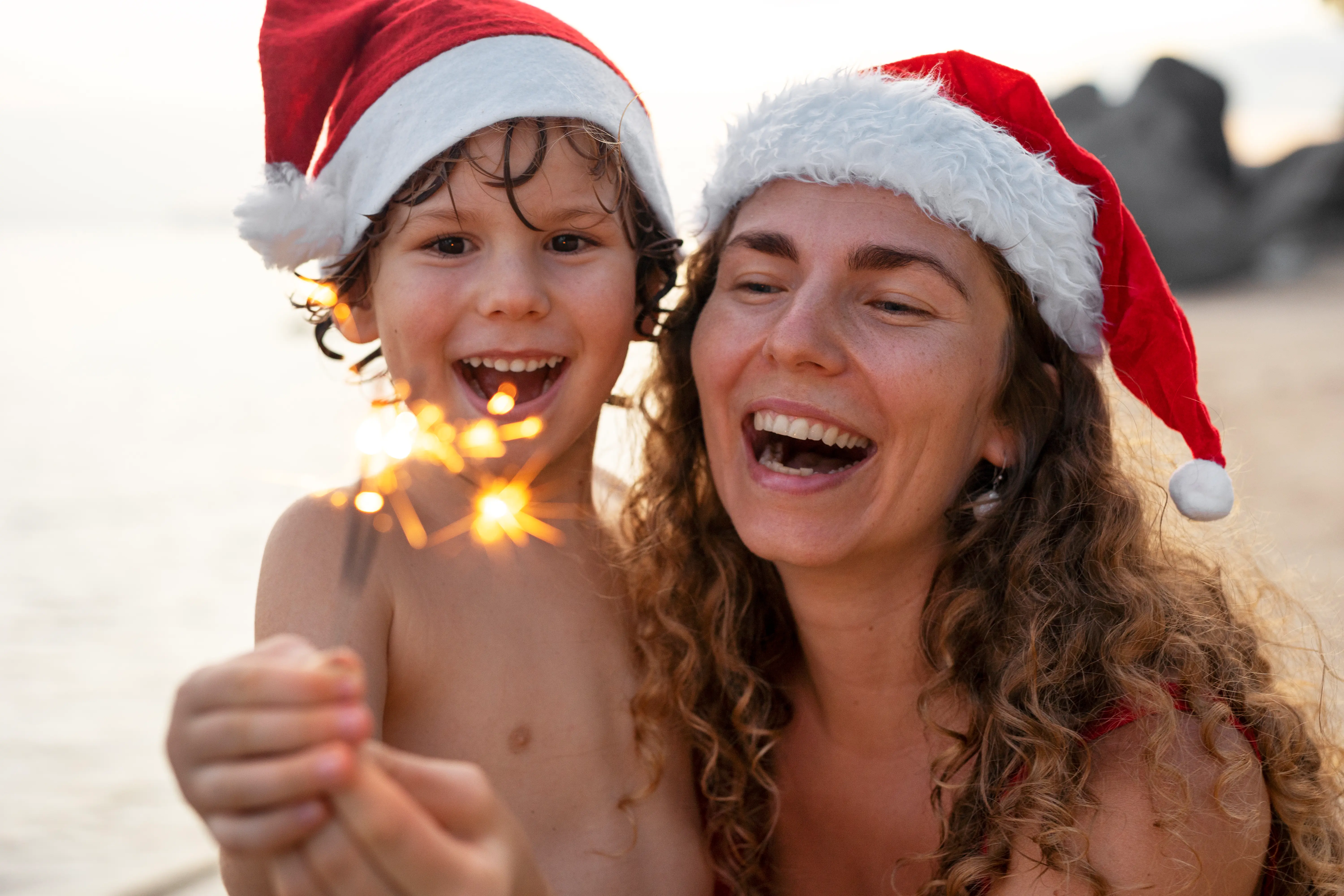 Warme Reiseziele im Dezember: Entfliehe dem Winter in dein Sommerglück Mutter und Kind tragen Weihnachtsmützen und halten eine brennende Wunderkerze am Strand, beide lachen fröhlich.