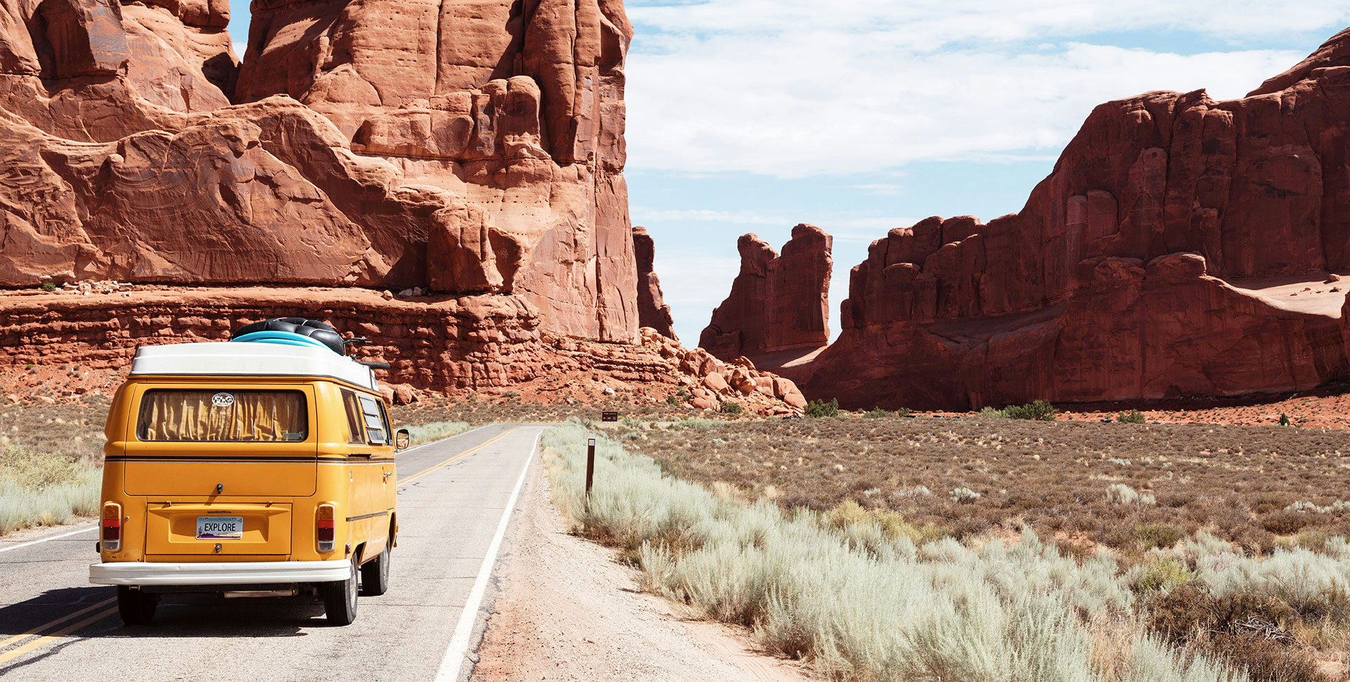 Dein Roadtrip durch die US-Nationalparks Ein gelber Campervan fährt durch eine imposante Landschaft mit roten Felsen und weitem Himmel.