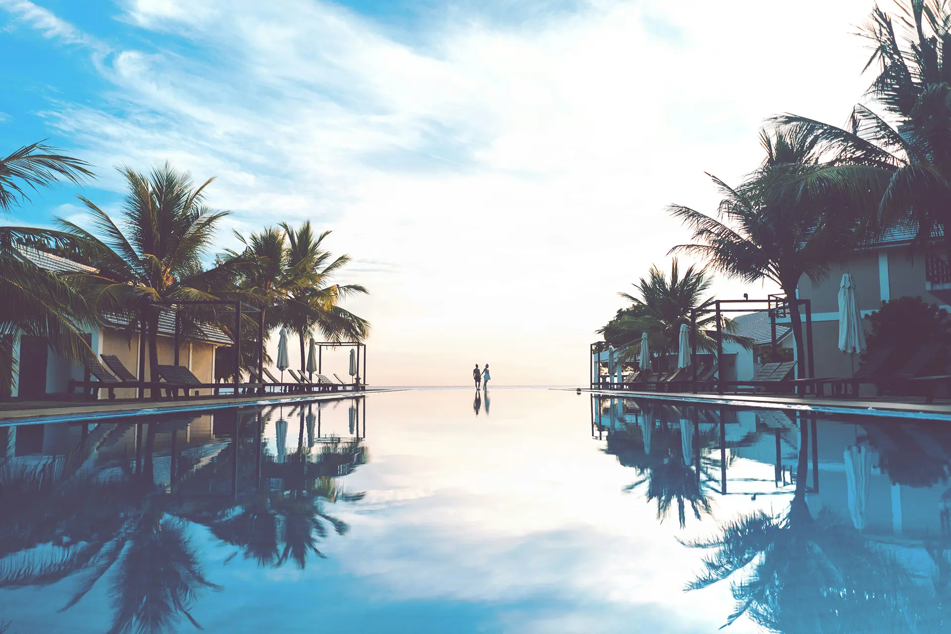 Airbnb & Co.: So findest Du die richtige Unterkunft Infinity-Pool in einem tropischen Resort mit Palmen, Liegen und zwei Menschen am Horizont bei Sonnenaufgang