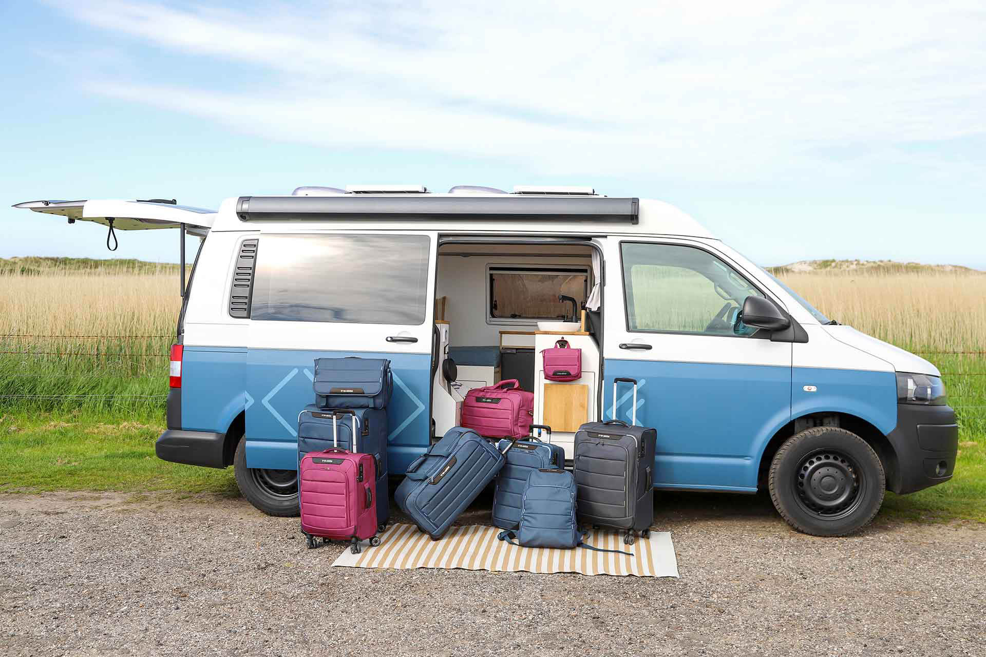 Campingbus mit geöffneten Türen und Reisekoffern auf einer Picknickdecke Ein blauer Camper steht auf einem Feldweg mit geöffneten Schiebetüren. Zahlreiche Koffer und Taschen in verschiedenen Farben befinden sich vor dem Fahrzeug auf einer gestreiften Picknickdecke.