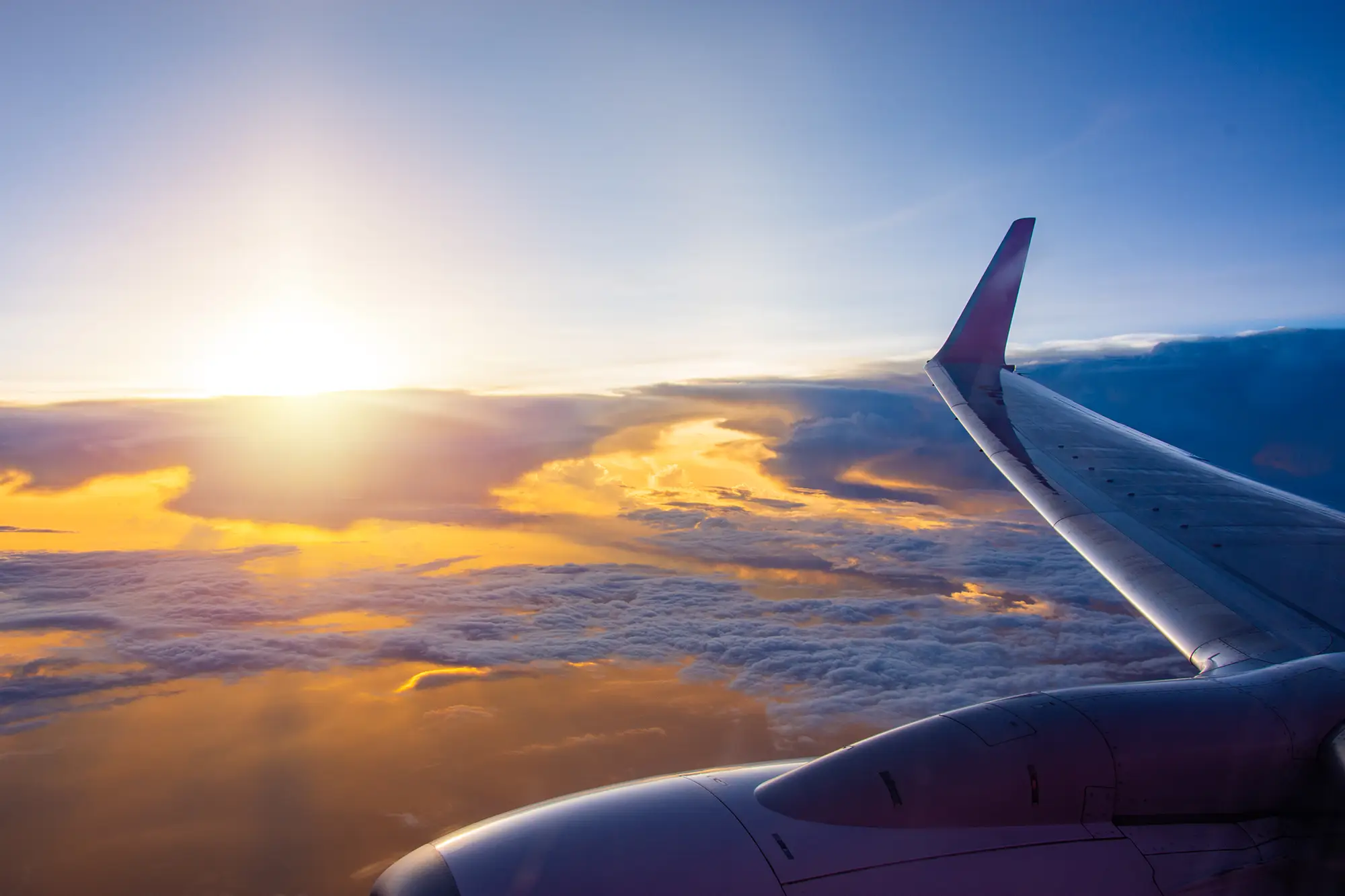 Tipps für lange Flugreisen – dein ultimativer Guide für Langstreckenflüge Flugzeugflügel im Sonnenaufgang, fotografiert aus dem Fenster über einer Wolkendecke