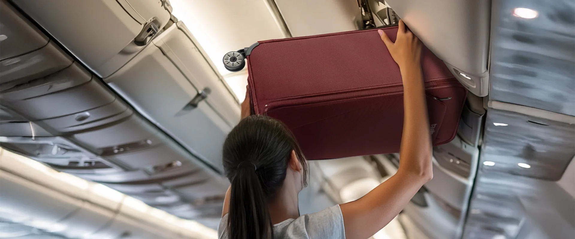 Handgepäckkoffer im Flugzeugfach – Praktischer Kabinenkoffer für Reisen Frau verstaut einen burgunderroten Handgepäckkoffer in das Gepäckfach eines Flugzeugs
