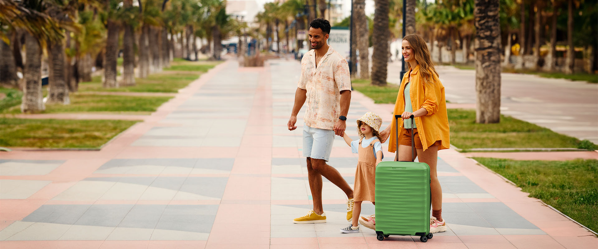 Familie im Urlaub mit grünem Koffer auf einer Promenade Eine fröhliche Familie mit einem grünen Koffer spaziert auf einer von Palmen gesäumten Promenade.