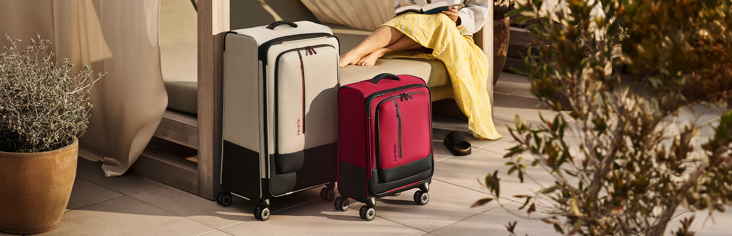 Two suitcases stand in a sunny outdoor area: one is beige, the other red, both with black accents. A person in a yellow dress sits in the background reading a book. Plants and a flower pot add to the relaxed atmosphere.