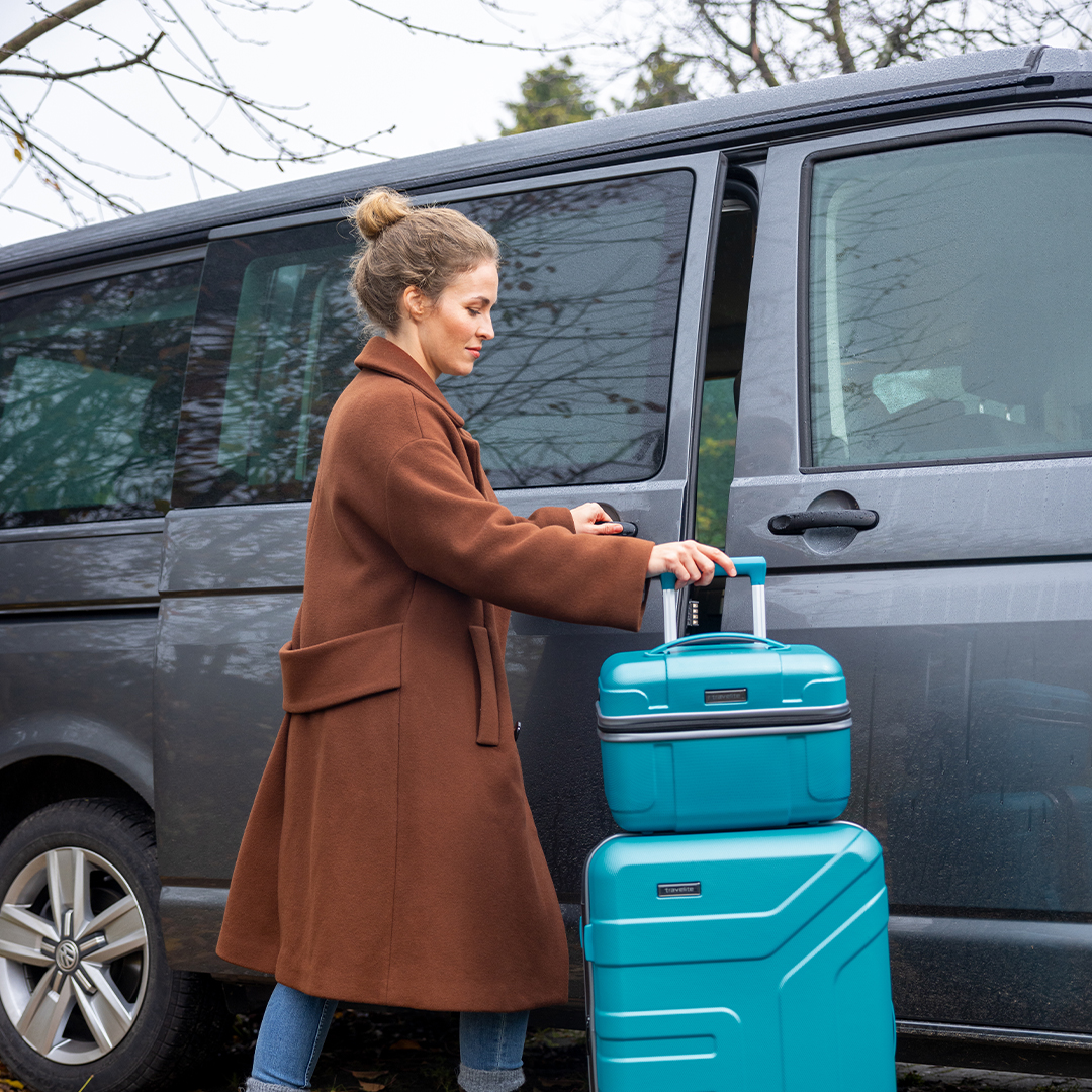 Frau mit braunem Mantel zieht blauen Koffer zu einem grauen Auto Eine Frau in einem braunen Mantel schiebt einen blauen Koffer zu einem geparkten grauen Van
