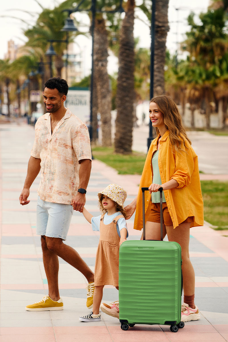 Familie beim Spaziergang mit grünem Koffer Eine lächelnde Familie mit kleinem Kind, die auf einer Promenade mit einem grünen Koffer spaziert. Palmen säumen den Weg.