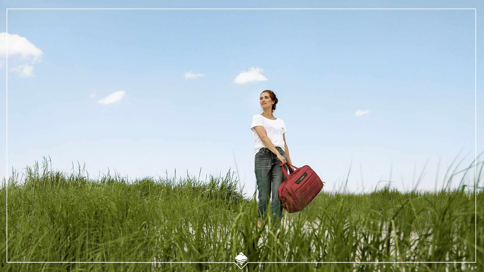 Frau mit roter Tasche im grünen Feld unter blauem Himmel Eine Frau steht in einem Feld aus grünem Gras und hält eine rote Tasche. Der Himmel ist klar und blau.