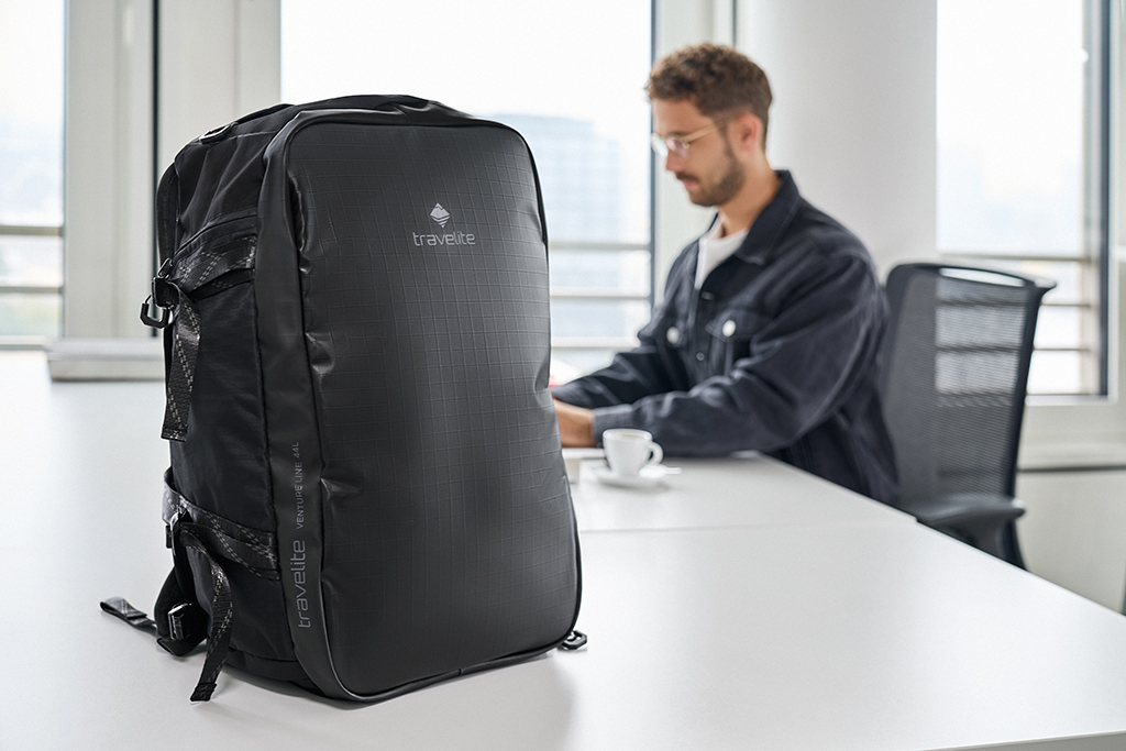 A black Travelite backpack stands on a white table. In the background, a blurred man can be seen at a desk with a cup of coffee. A building is recognisable through a window.