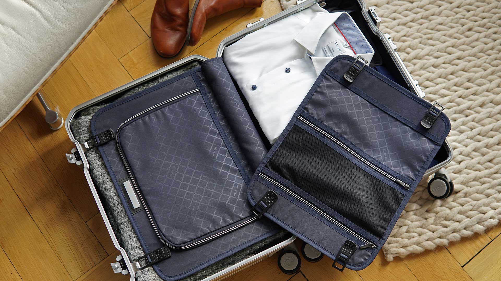 Ein geöffneter Koffer auf einem Holzboden, der Hemden und Kleidung enthält, neben einem Teppich und Lederschuhen.