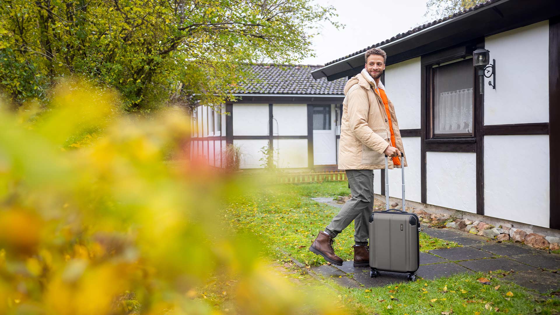 Mann zieht Koffer in Herbstlandschaft vor einem Fachwerkhaus Ein Mann in warmer Kleidung zieht einen Rollkoffer auf einem gepflasterten Weg entlang, während im Hintergrund ein Fachwerkhaus und herbstliche Bäume zu sehen sind.