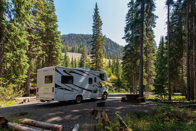 Holidays with a caravan: enjoy freedom on four wheels Ein Wohnmobil steht auf einem Wald-Campingplatz neben einer Picknickbank, umgeben von hohen Bäumen und Bergen im Hintergrund