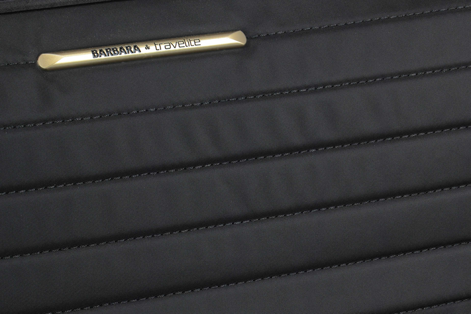 Detailansicht eines schwarzen Koffers mit gestepptem Stoff und Markenlabel 'BARBARA & travelite' Nahaufnahme des gesteppten schwarzen Stoffes eines Koffers mit einem Metalllabel der Marke 'BARBARA & travelite'.