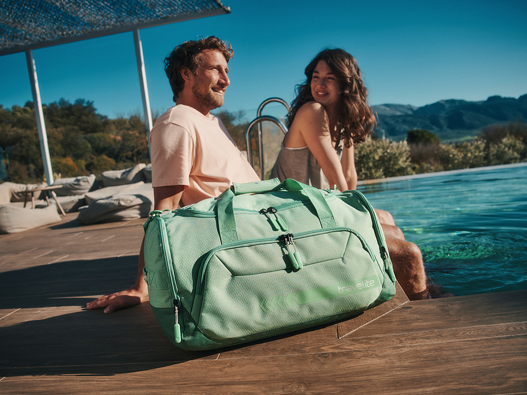 Ein Paar sitzt lächelnd am Pool, mit den Füßen im Wasser. Im Vordergrund steht eine hellgrüne Reisetasche der Marke Travelite auf einem hölzernen Beckenrand. Im Hintergrund sind Bäume und Berge unter einem blauen Himmel sichtbar.