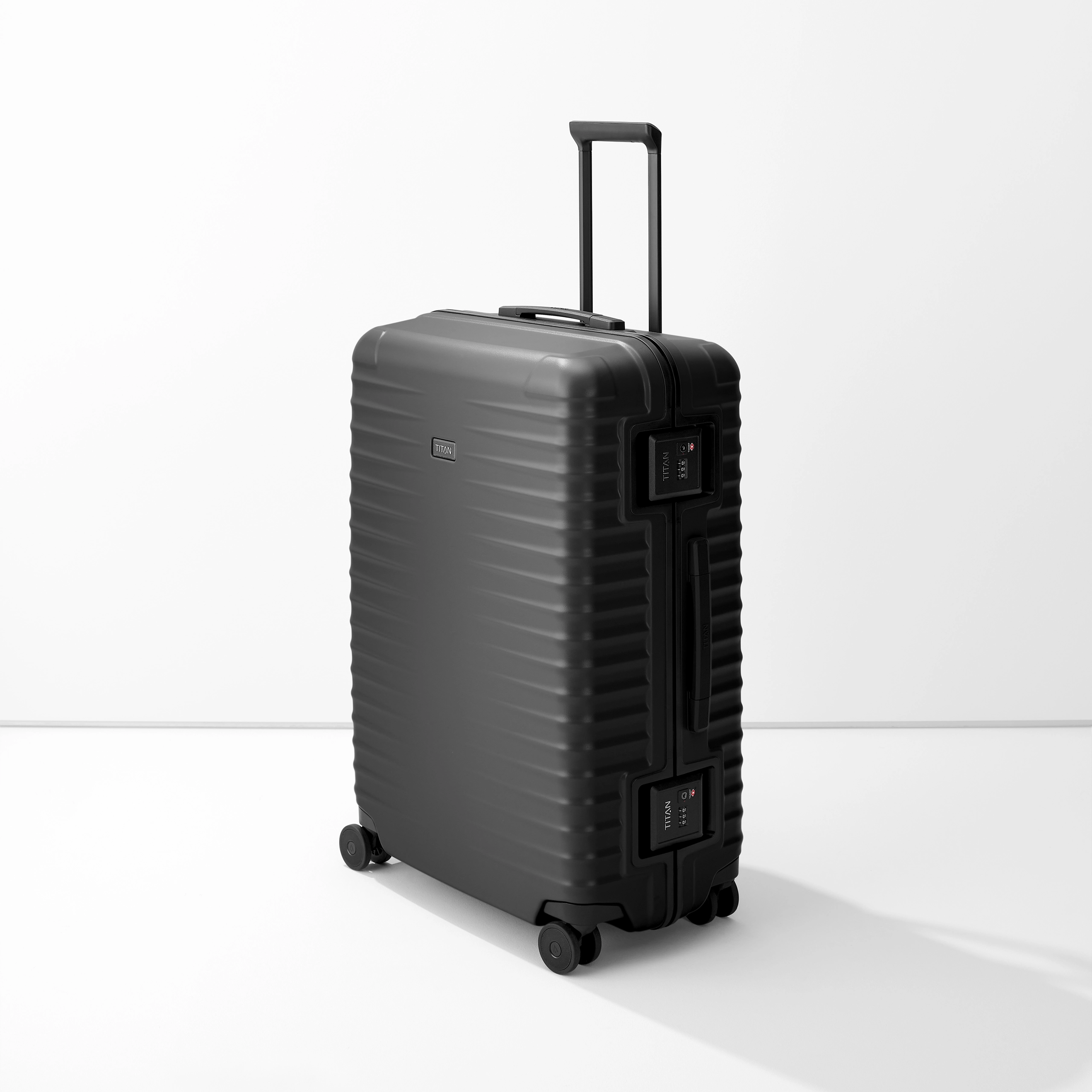 Un trolley L nero TITAN poggia su una base bianca. La valigia ha una superficie a coste e una maniglia estensibile. Sul lato sono visibili due lucchetti TSA. La valigia poggia su quattro ruote doppie.