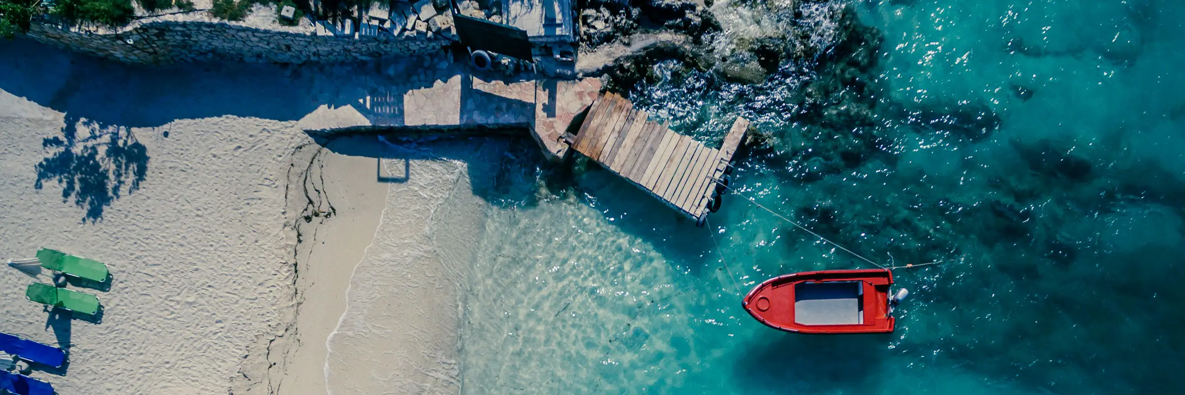 Insider tips for holidays in August: the best destinations for sun, nature & culture Luftaufnahme eines idyllischen Strandes mit Holzsteg und rotem Boot im klaren, türkisblauen Wasser